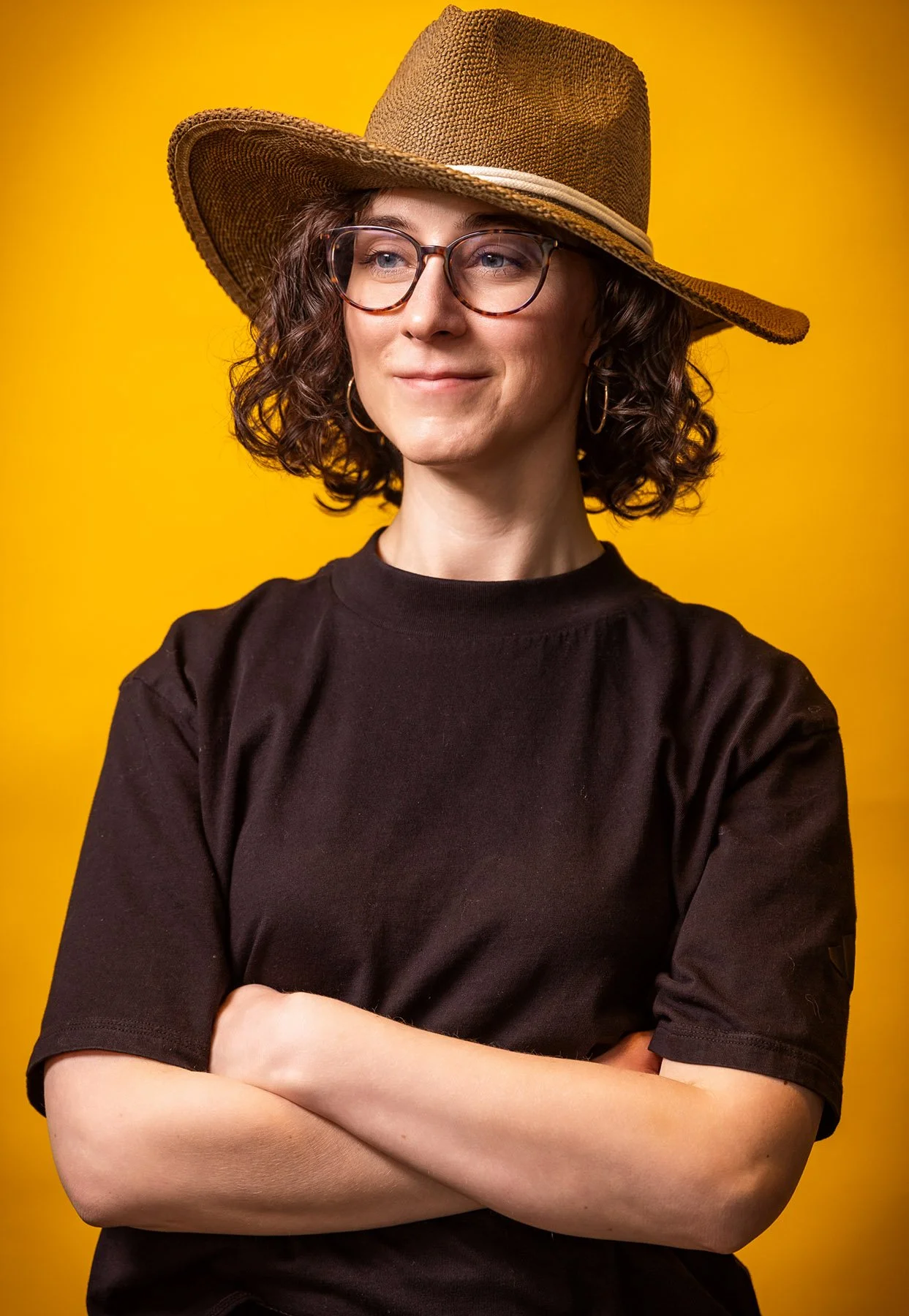 Woman in a straw hat and black tee with arms crossed on a golden yellow backdrop