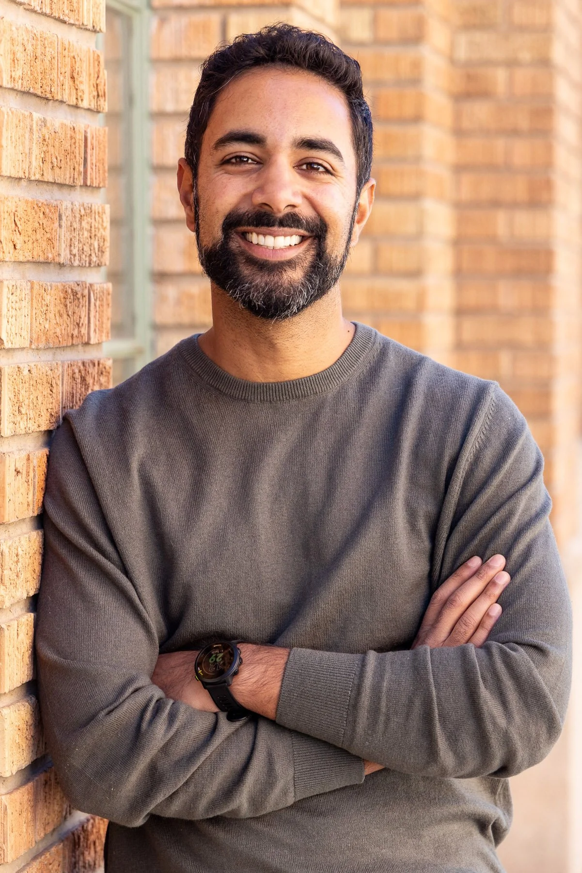 Smiling man in an olive sweater with arms crossed against a brick wall