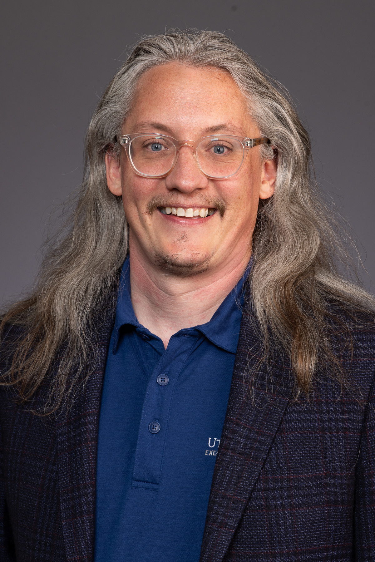 Executive with long grey hair, clear glasses and a plaid blazer over a blue polo