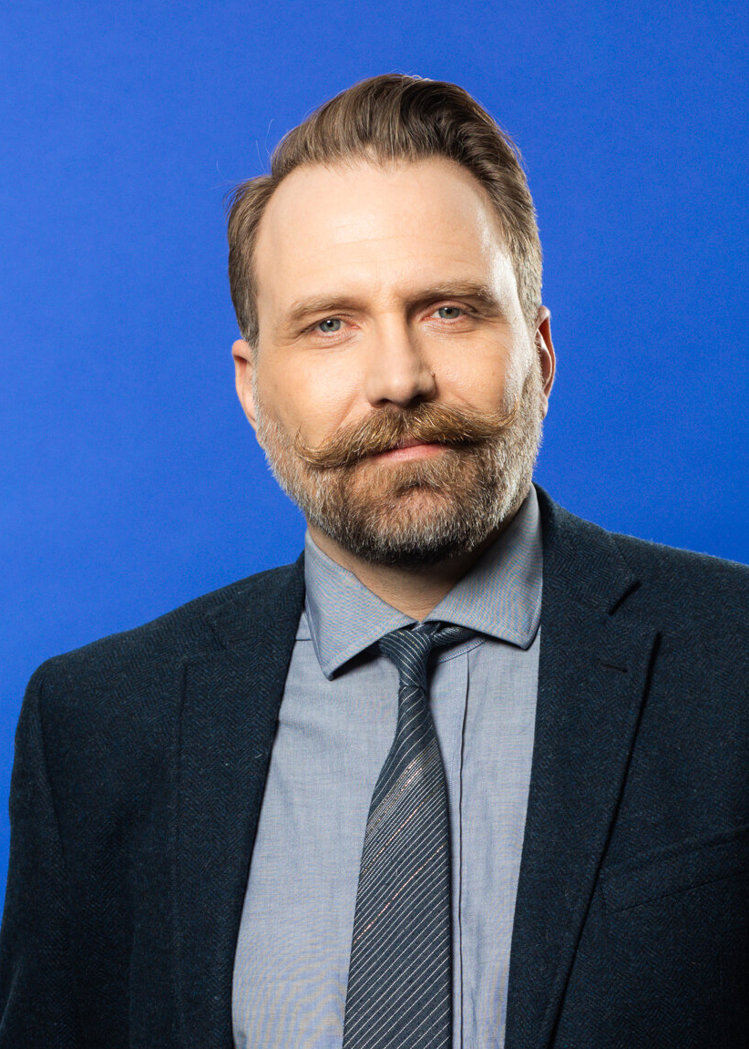 Bearded man in a charcoal suit and tie on a cobalt blue backdrop